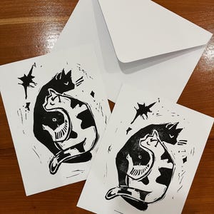 Op de afbeelding: Twee rechthoekige witte wenskaarten met een zwarte linoleumsnede van twee katten die in een yin-yangvorm tegen elkaar aan liggen, vergezeld van een bijpassende witte envelop. Het ontwerp bevat gestileerde stervormen.