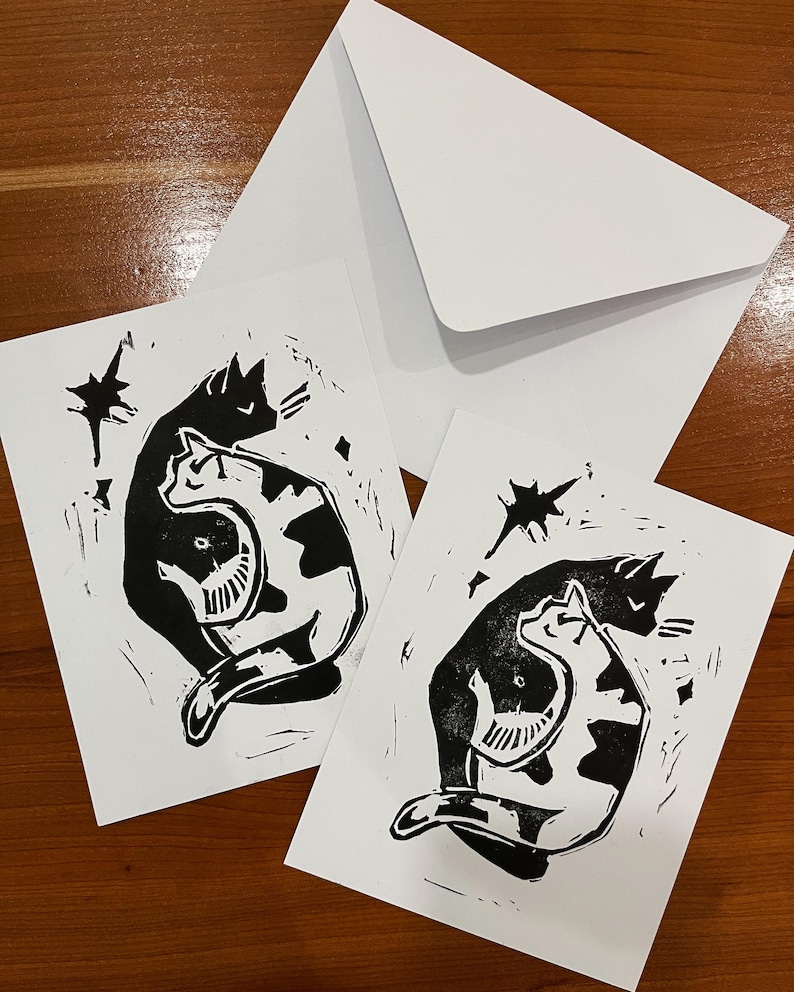 Op de afbeelding: Twee rechthoekige witte wenskaarten met een zwarte linoleumsnede van twee katten die in een yin-yangvorm tegen elkaar aan liggen, vergezeld van een bijpassende witte envelop. Het ontwerp bevat gestileerde stervormen.