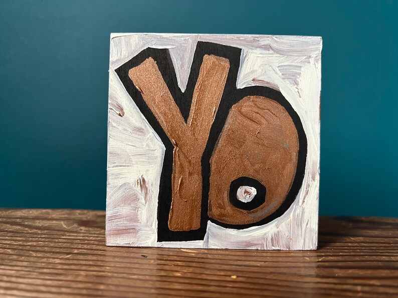 May include: A wooden block painted white with a gold and black stylised letter "Y" and a gold and black stylised letter "O".
