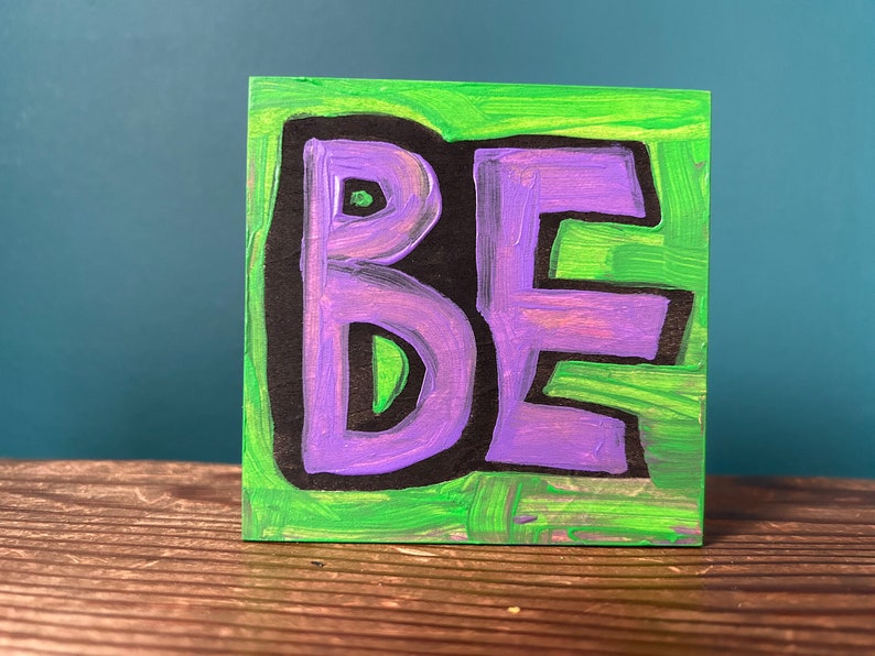May include: A small, square, hand-painted artwork featuring the word "BE" in purple, outlined in black, against a green background. The letters are bold and stylized, with a slightly textured appearance. The artwork is displayed on a wooden surface.