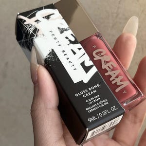 May include: A Fenty Beauty Gloss Bomb Cream lip gloss in a clear tube with a metallic cap, next to a black box. The box has white text and graphics, including "Gloss Bomb Cream" and "Color Drip Lip Cream". The lip gloss is a reddish-pink color.