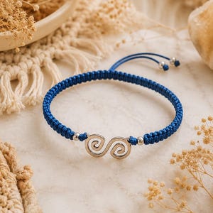 Puede incluir: Una pulsera de cordón trenzado azul con un dije central de metal plateado en forma de dos espirales conectadas. La pulsera incluye dos pequeñas cuentas plateadas a los lados del dije y cordones ajustables con terminales plateados.