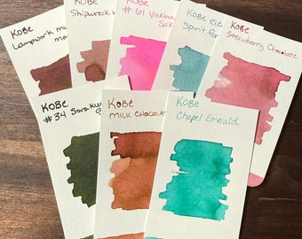 May include: Eight rectangular white paper swatches featuring ink samples in various colors including dark brown, muted rose, bright pink, teal, strawberry red, forest green, light brown, and emerald green, each labeled with Kobe ink names.