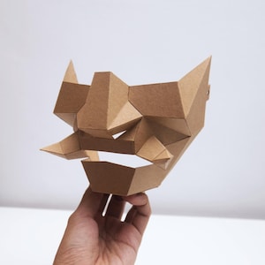 May include: A geometric, brown paper mask with sharp angles and points. The mask has a stylized, angular design, with a prominent brow, nose, and mouth area. The mask is held in a hand against a white background.