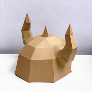 May include: A brown papercraft Viking helmet with two curved horns and a central spike. The helmet is constructed from geometric shapes, creating a polygonal design. The craft is set against a white background.