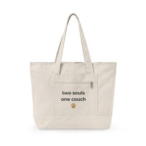 May include: Cream-colored tote bag with a front pocket and zipper closure. The text "two souls one couch" is printed on the front, along with a paw print graphic. The bag has two long handles.
