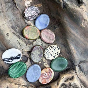 May include: A collection of eleven smooth, oval-shaped polished stones in various colors and patterns, including green malachite with stripes, white with black veins, spotted dalmatian jasper, and solid blue, pink, and green mineral specimens.