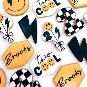 May include: A collection of decorated sugar cookies featuring yellow smiley faces with lightning bolts, black lightning bolt shapes, white cookies with the text two cool, black and white checkered number twos, and yellow hexagons with the name Brooks.