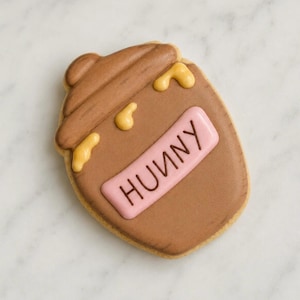 May include: A brown cookie shaped like a honey pot with the word "HUNNY" on a pink rectangle. The cookie has yellow honey drips and is on a white surface.