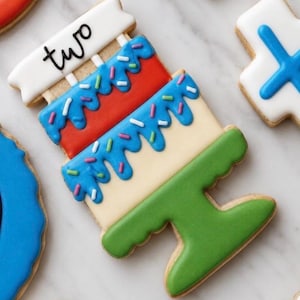 Könnte beinhalten: Ein dekorierter Keks in Form einer zweistöckigen Torte. Die oberste Schicht ist rot, die mittlere Schicht ist blau und die untere Schicht ist beige. Der Kuchen ist mit blauer Glasur, Streuseln und dem Wort "two" auf einem weißen Banner verziert.