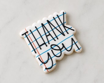 May include: A rectangular sugar cookie decorated with white icing to resemble a piece of notebook paper. The cookie features horizontal blue lines, a vertical red margin line, and the words THANK YOU written in bold black icing.