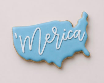 Può includere: Un biscotto a forma di Stati Uniti contigui, ricoperto da una glassa liscia color azzurro. La parola 'Merica è scritta al centro in glassa bianca corsiva, con l'apostrofo posizionato prima della lettera M.