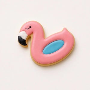 May include: A flamingo-shaped cookie decorated with smooth pink icing, featuring a white and black beak, a closed eye detail, and a central oval of light blue icing. The cookie has a visible tan-colored edge and a glossy, finished surface.
