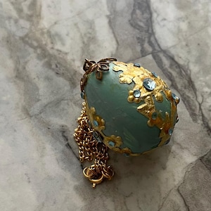 Könnte beinhalten: Ein hellblauer, eiförmiger Anhänger mit filigranen goldfarbenen Metallauflagen und kleinen, eingelassenen hellblauen Kristallen. Das Schmuckstück ist an einer zarten goldfarbenen Metallkette mit einem runden Verschluss befestigt.
