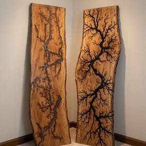 May include: Two tall, rectangular live-edge wood slabs standing upright. Each slab features a prominent, dark, branching fractal pattern burned into the natural light brown wood grain, creating a complex, tree-like design across the surface.