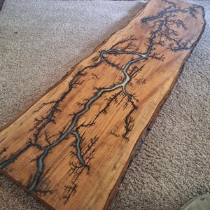 May include: A rectangular wooden board with a natural live edge, featuring a dark, intricate fractal burn pattern resembling lightning or tree branches that runs along the center, with a smooth, light brown wood grain finish throughout the surface.