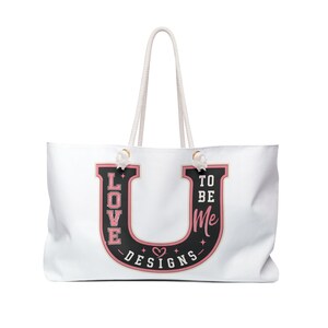 May include: A white tote bag with rope handles featuring a large black letter U graphic. The letter contains the pink text LOVE, TO BE, and ME, along with the white text DESIGNS and a small heart icon. The graphic is outlined in pink.