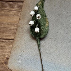 以下が含まれることがあります： 刺繍された緑の葉と、4つの小さな白い鈴蘭の花があしらわれたラペルピン。デザインは細い金属製のピンに取り付けられており、葉の付け根部分は緑色の糸で巻かれています。