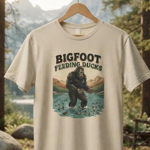 May include: A beige short-sleeve t-shirt featuring a graphic illustration of a dark brown Bigfoot standing in a lake surrounded by ducks. The text above the illustration reads BIGFOOT FEEDING DUCKS in a dark brown, bold, arched font style.