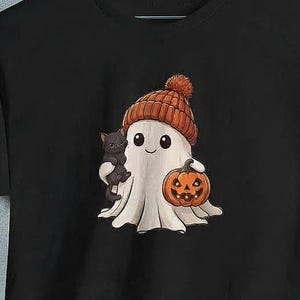 Può includere: Una t-shirt nera a maniche corte con una stampa grafica di un fantasma bianco che indossa un berretto di lana arancione. Il fantasma tiene un piccolo gatto nero in un braccio e una zucca arancione intagliata nell'altra mano.