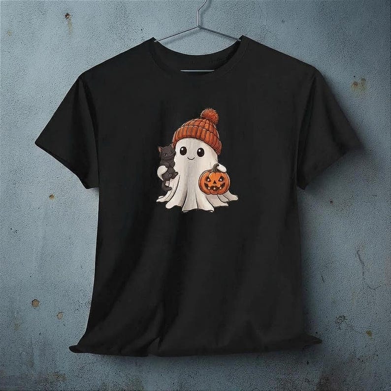 Può includere: Una t-shirt nera a maniche corte con una stampa grafica di un fantasma bianco che indossa un berretto di lana arancione. Il fantasma tiene un piccolo gatto nero in un braccio e una zucca arancione intagliata nell'altra mano.