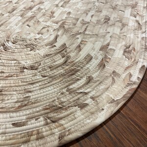 May include: An oval-shaped rug constructed from coiled fabric strips. The material features a repeating pattern of soft beige, cream, and muted brown tones. The coils are joined together with visible, tight, light-colored zigzag stitching.
