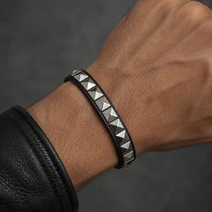May include: A black leather bracelet featuring a single row of silver-toned metal pyramid studs. The bracelet is designed with a smooth band and a series of evenly spaced, raised square-based studs that run along the center of the leather strap.