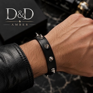 May include: A black leather bracelet featuring four evenly spaced silver-toned metal spikes. The text D&D AMBER is visible in the upper left corner. The bracelet is worn on a wrist, with a black leather jacket sleeve visible at the edge of the frame.