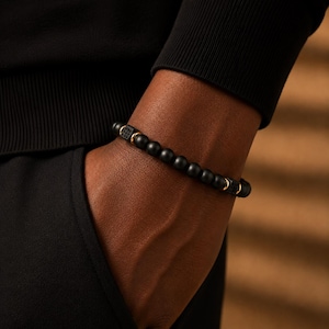 May include: A beaded bracelet featuring smooth matte black spherical beads interspersed with polished gold-tone metal rings and two square black textured accent beads. The design consists of a repeating pattern of dark beads and metallic gold spacers.