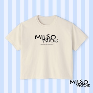 May include: Cream-colored t-shirt with the text "MILSO PRIDE" in black, with the tagline "ONE MILITARY WIFE TO ANOTHER" below. The shirt is displayed against a light blue and white striped background.