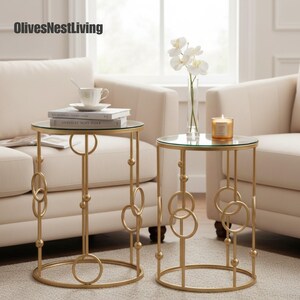 May include: Two gold side tables with glass tops. The tables have a circular base and a decorative design of interlocking circles and spheres. One table holds books and a teacup, the other a candle and a vase of flowers.