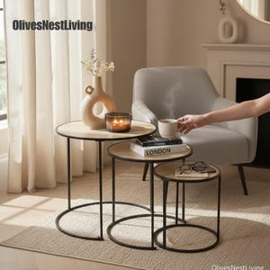 May include: Three round nesting tables with light wood surfaces and black metal frames. A vase, candle, and books are on the tables. The text "OlivesNestLiving" is in the top left corner.