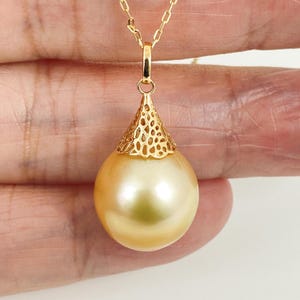 May include: A golden South Sea pearl pendant suspended from a delicate gold chain. The pearl is round and lustrous, topped with an intricate, openwork gold filigree cap that connects to a small bail, creating a refined and elegant jewelry piece.