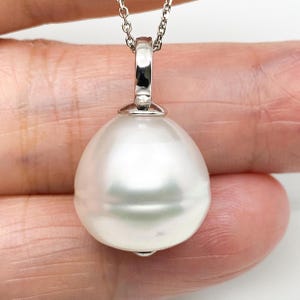May include: A large, lustrous white pearl pendant suspended from a polished silver-toned metal bail and a delicate chain. The pearl has a smooth, rounded shape with a reflective surface, showcasing a bright, clean appearance and a classic design.