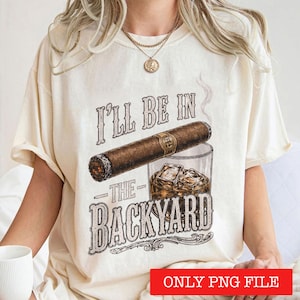 May include: A cream-colored short-sleeve t-shirt featuring a graphic of a cigar resting on a glass of whiskey with ice cubes. The text "I'LL BE IN THE BACKYARD" is printed in a distressed font above and below the cigar and glass illustration.