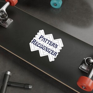 Op de afbeelding: Een gestanste sticker met een lichtgrijze visgraatpatroon achtergrond en de donkerblauwe tekst Pattern Recognizer gedrukt in een vet, schreefloos lettertype in het midden, aangebracht op een zwart skateboard deck.