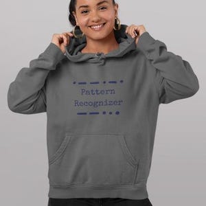 May include: A charcoal gray hooded sweatshirt featuring a front kangaroo pocket and the text Pattern Recognizer printed in dark blue on the chest, accompanied by decorative Morse code-style dots and dashes above and below the lettering.