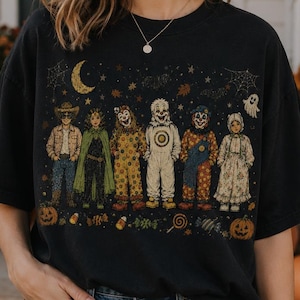 May include: A black short-sleeve t-shirt featuring a graphic print of six figures in various Halloween costumes, including clowns and a ghost, surrounded by small icons like a crescent moon, spiderwebs, candy, and pumpkins at the bottom.