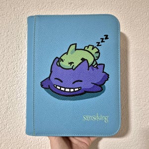 Peut inclure: Un classeur bleu clair texturé avec une illustration d'une créature violette au large sourire et d'une petite créature verte endormie sur elle. Le texte "sanseking" est imprimé dans le coin inférieur droit en police cursive vert clair.