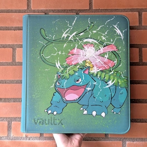Op de afbeelding: Een talingkleurige verzamelkaartenmap met een grafische illustratie van het wezen Venusaur met een roze bloem op zijn rug. Het ontwerp bevat witte bliksemachtige effecten en de tekst vaultx gedrukt in de linkerbenedenhoek.