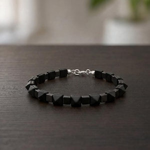 Pode incluir: Uma pulseira com alternância de contas quadradas em preto mate e tachas pretas em forma de pirâmide, ligadas por um fecho de mosquetão em metal prateado. O design consiste num padrão repetitivo de cubos de faces planas e formas geométricas.