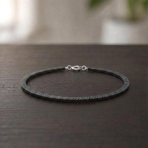 May include: A dark gray, chevron-patterned beaded necklace or bracelet featuring a series of small, interlocking geometric segments. The piece is finished with a polished silver-toned metal lobster claw clasp and circular jump ring connector.