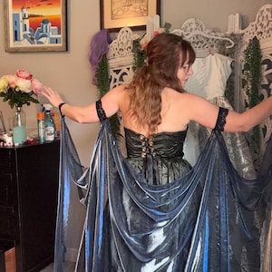 May include: A dark green and black corset-style dress with a flowing, iridescent blue fabric attached to the sleeves. The dress has a lace-up back and black lace accents. The image shows a woman wearing the dress.