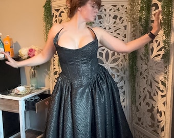 May include: A woman wearing a black, sparkly, strapless gown with a fitted bodice and a full skirt. She is standing in front of a white, carved screen with green plants.