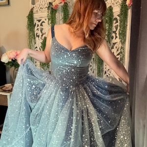 Op de afbeelding: Een formele jurk met een sweetheart halslijn en een volle rok. De bodice is van zilverblauw fluweel en de rok is van gelaagde tule, beide versierd met sprankelende stervormige versieringen. De jurk heeft een donkerblauwe band.