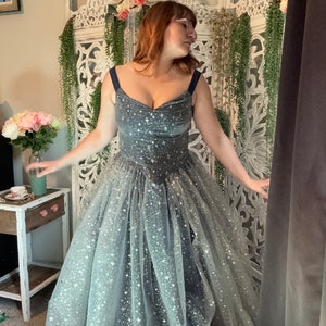 Op de afbeelding: Een donkergrijze galajurk met een sweetheart halslijn en een zwierige tule rok. De jurk is versierd met zilveren stervormige glitter. De bandjes zijn marineblauw. De jurk is geschikt voor een gala of een speciale gelegenheid.