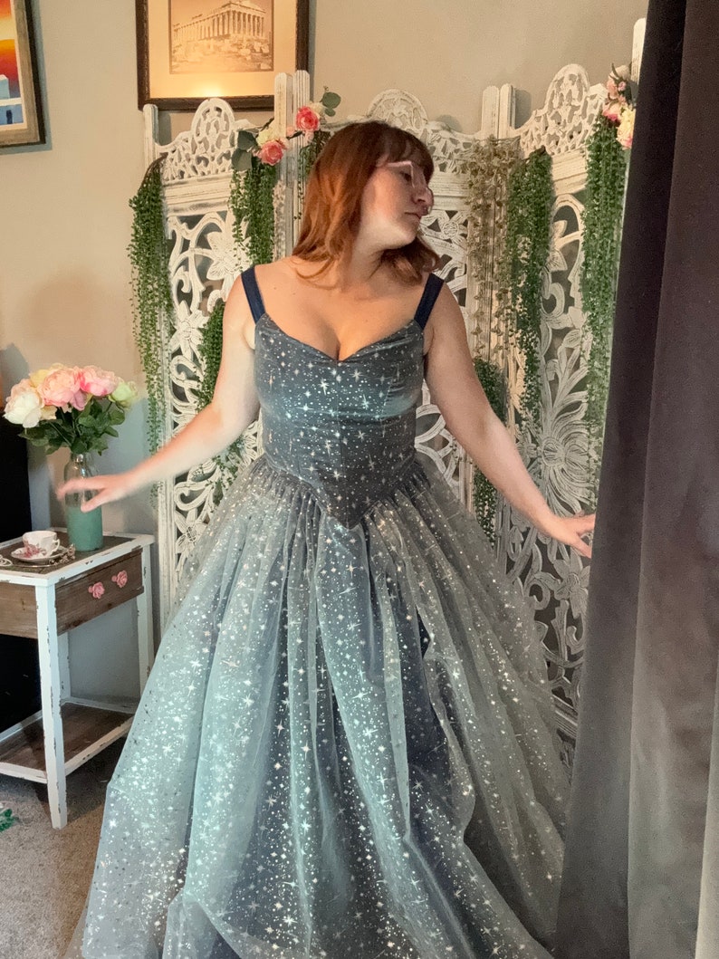 Op de afbeelding: Een donkergrijze galajurk met een sweetheart halslijn en een zwierige tule rok. De jurk is versierd met zilveren stervormige glitter. De bandjes zijn marineblauw. De jurk is geschikt voor een gala of een speciale gelegenheid.