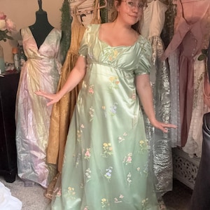 Könnte beinhalten: Ein hellgrünes Satin-Kleid mit kurzen Ärmeln und fließendem Rock. Das Kleid ist mit gestickten floralen Details in Rosa, Gelb und Blau verziert. Das Kleid hat eine hohe Taille und einen Sweetheart-Ausschnitt, geeignet für formelle Anlässe.