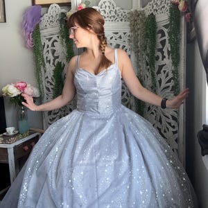 May include: A woman wearing a light blue tulle ball gown with silver glitter stars. The gown has a sweetheart neckline and spaghetti straps.
