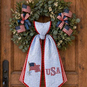 May include: A decorative wreath featuring green eucalyptus leaves, small white flowers, and four miniature American flags. A light blue and white gingham fabric bow with red trim hangs from the center, displaying an embroidered flag and USA text.